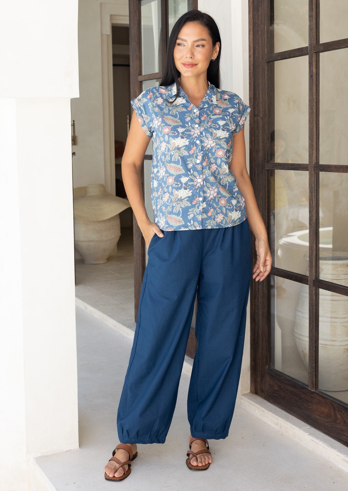 Woman stands wearing a dark blue 100% cotton wide leg pants with pleats at the ankles, with hand in pocket.