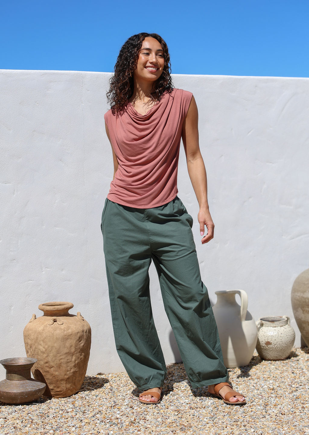 Woman wearing a pink sleeveless top and green pants standing outdoors with a white wall and blue sky in the background.