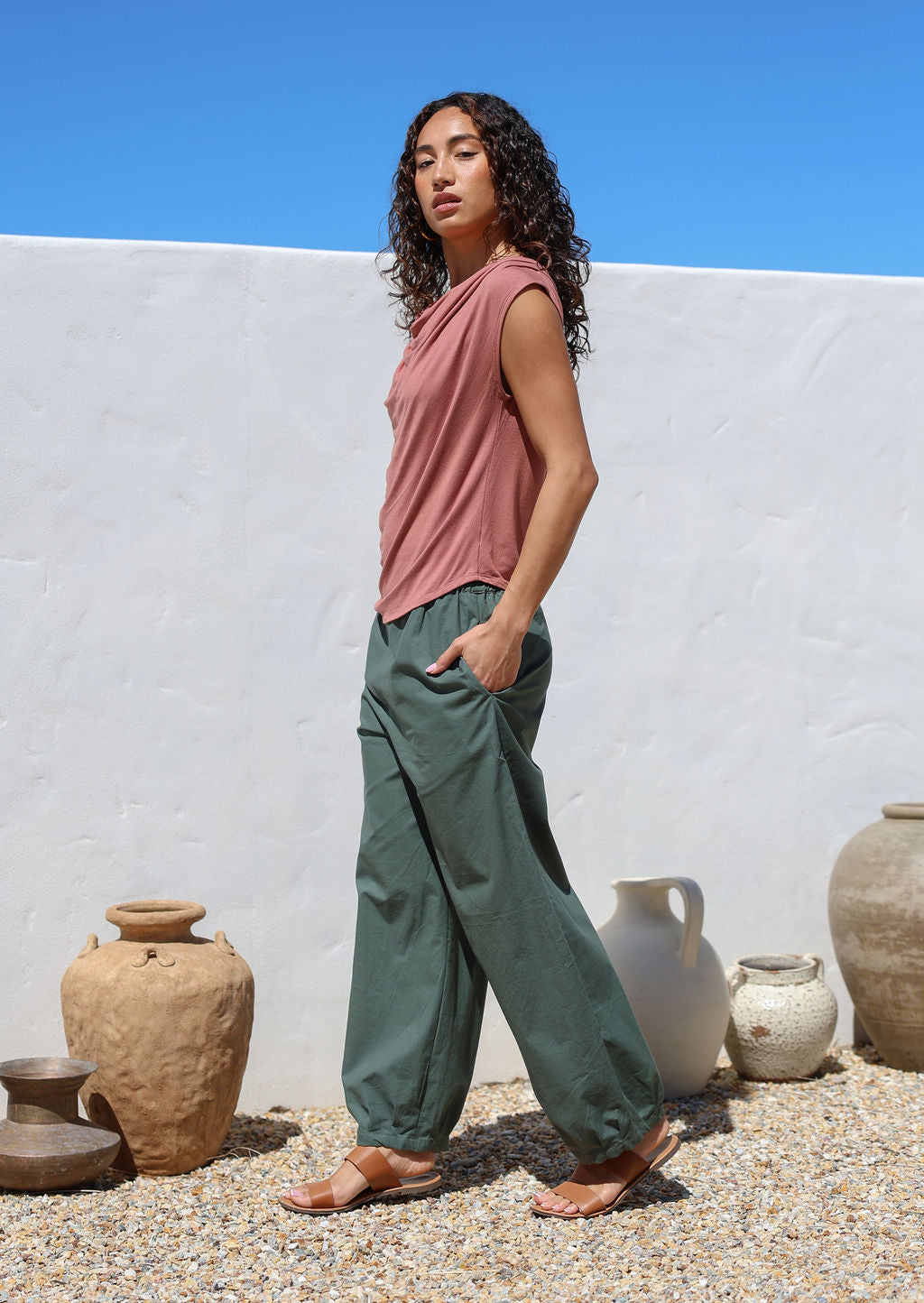 Woman in a pink top and green pants standing side on with hands in pockets
