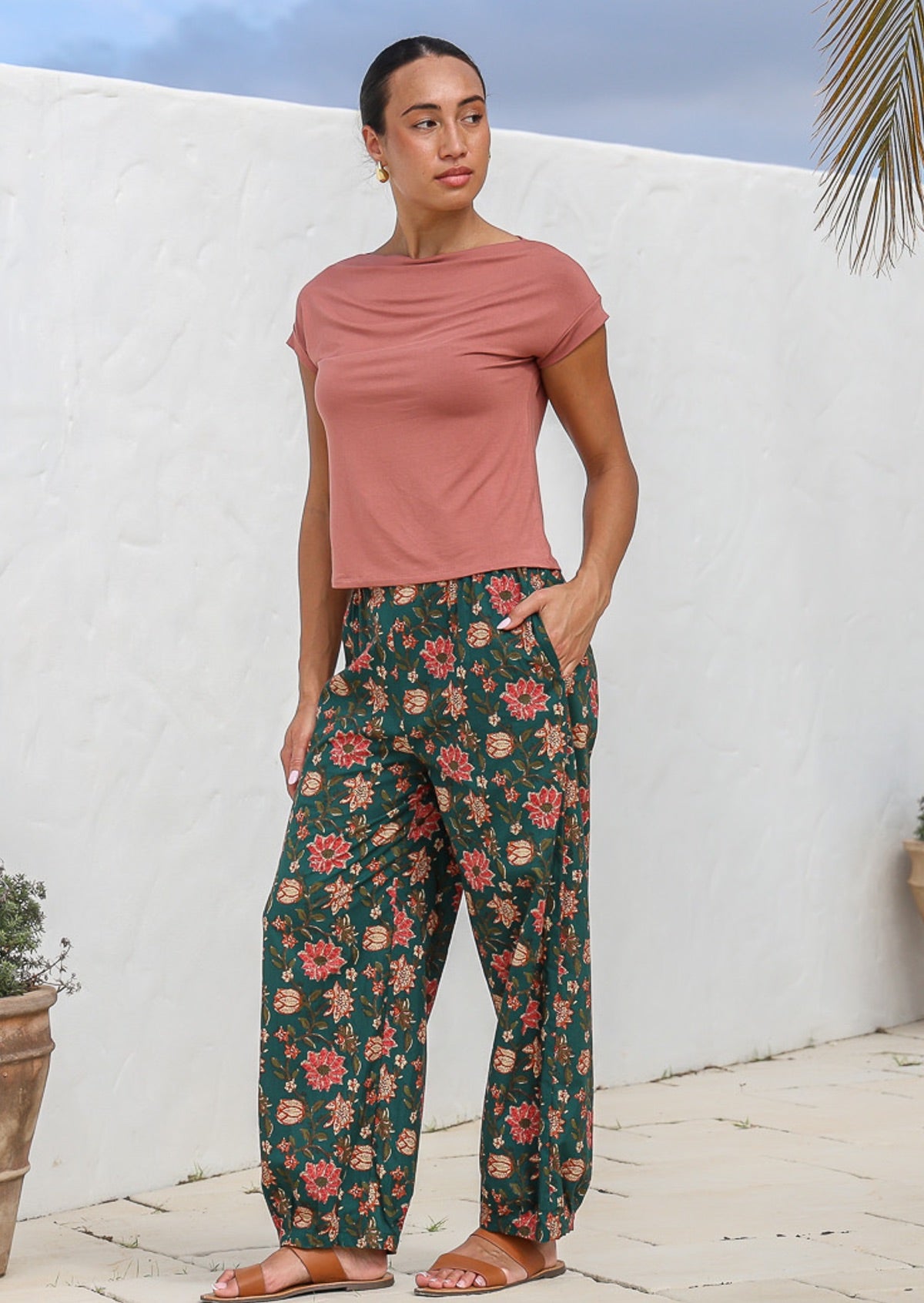 Model stands with a hand in pocket wearing 100% cotton floral print pants in deep green.