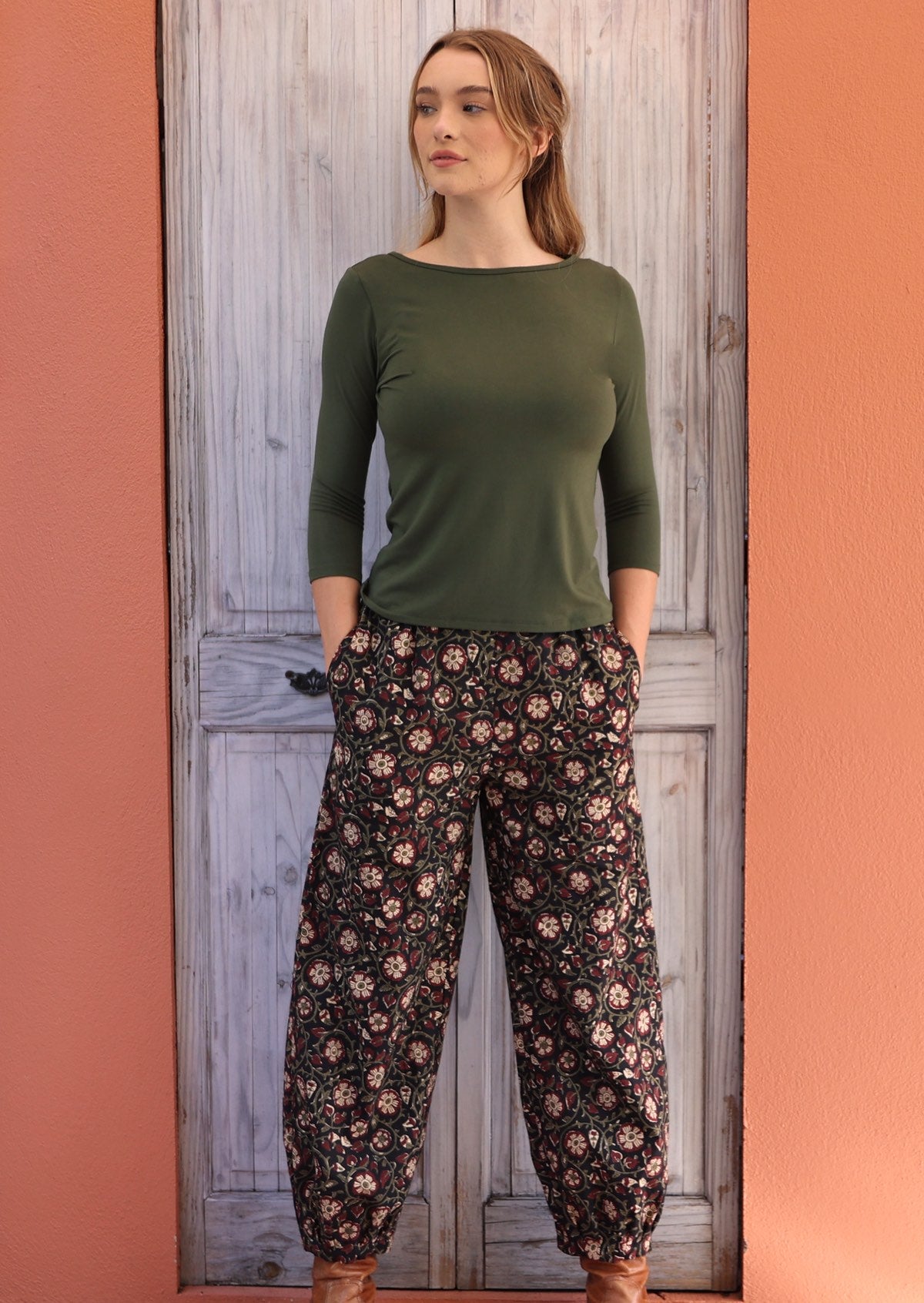 Woman standing in front of white door with hands in pockets wearing black based floral Indian 100% cotton pants
