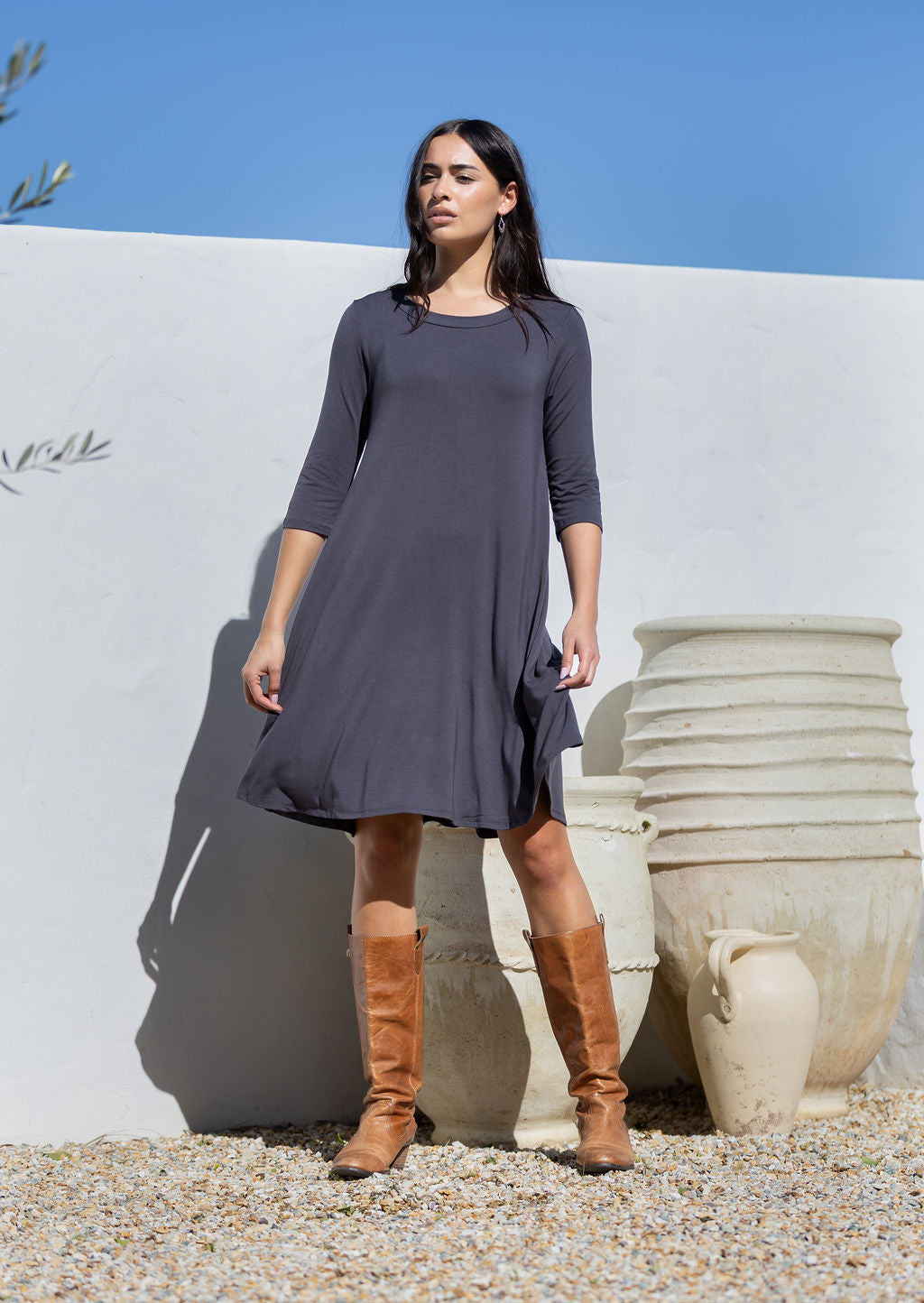 Woman wearing a dark gray jersey dress and brown knee-high boots standing against a white wall with clay pots.