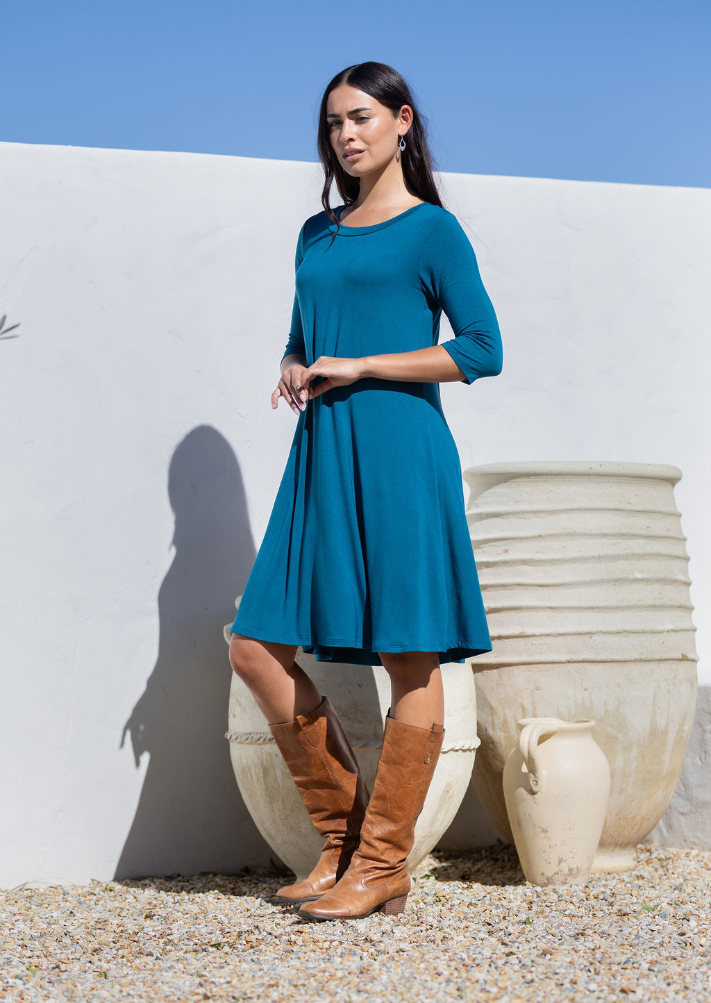 Woman in a teal rayon jersey dress and brown boots standing against a white wall with blue sky.