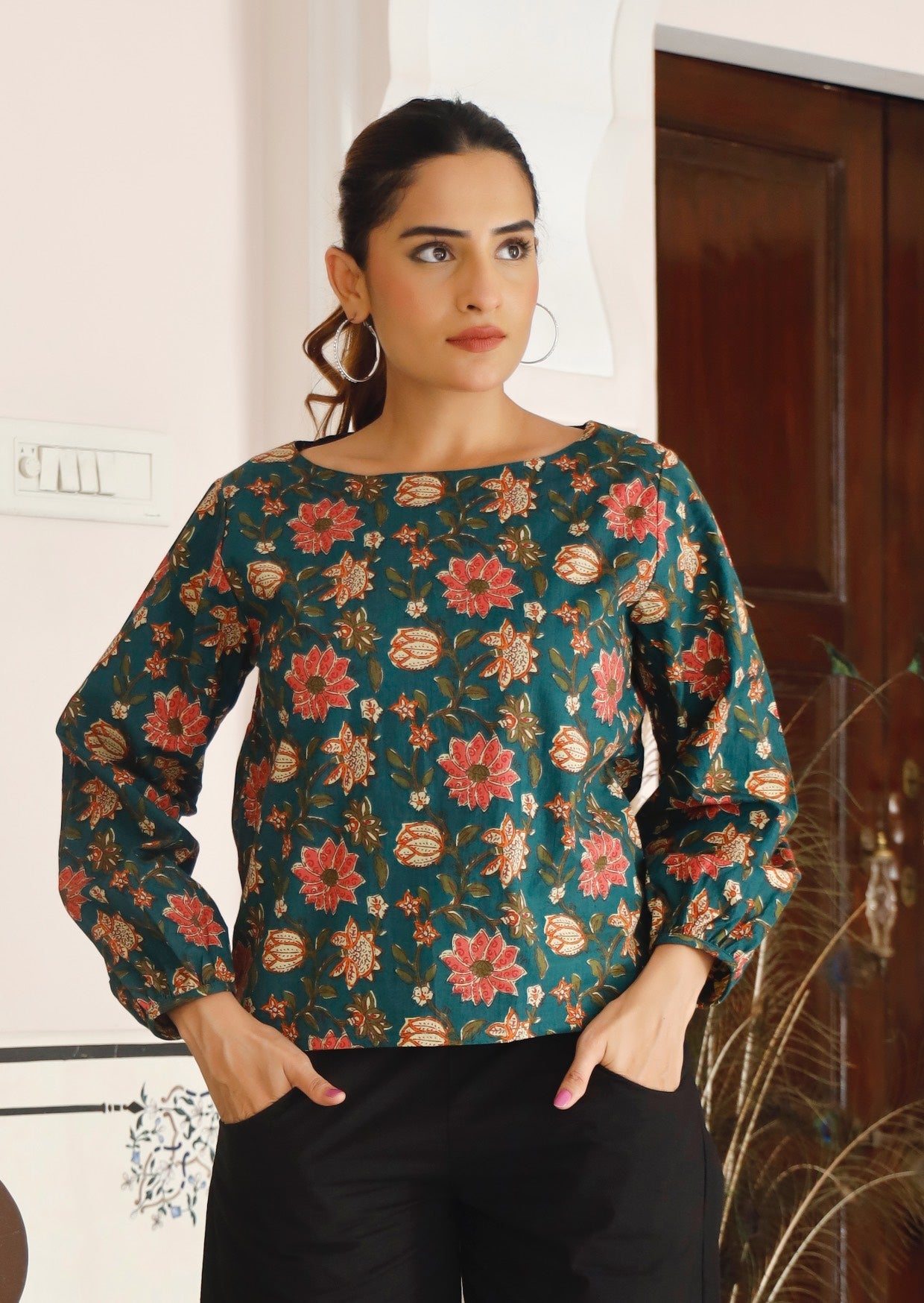 Woman with hands in pants pocket wears loose fit cotton top with wide round neckline and bishop sleeves.