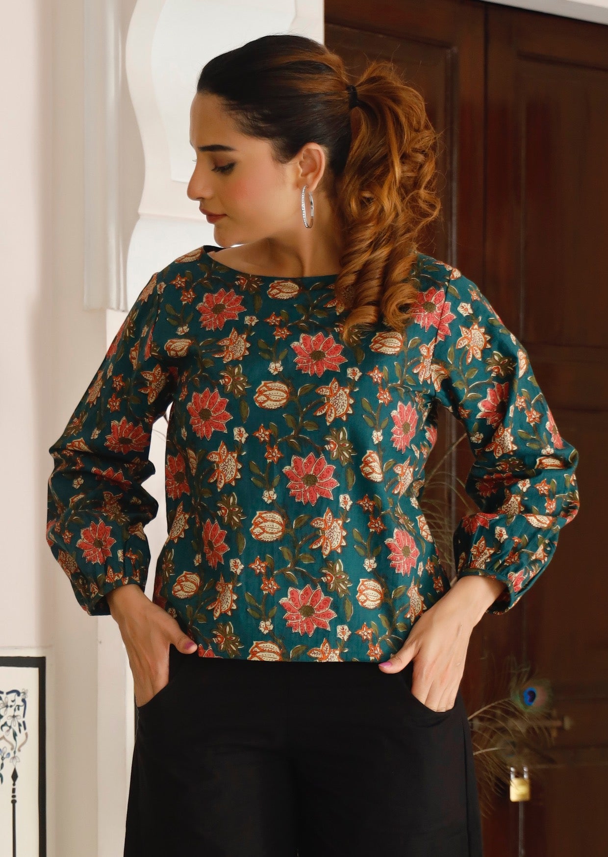 Woman wearing deep green based floral printed top with boat neckline and bishop sleeves.