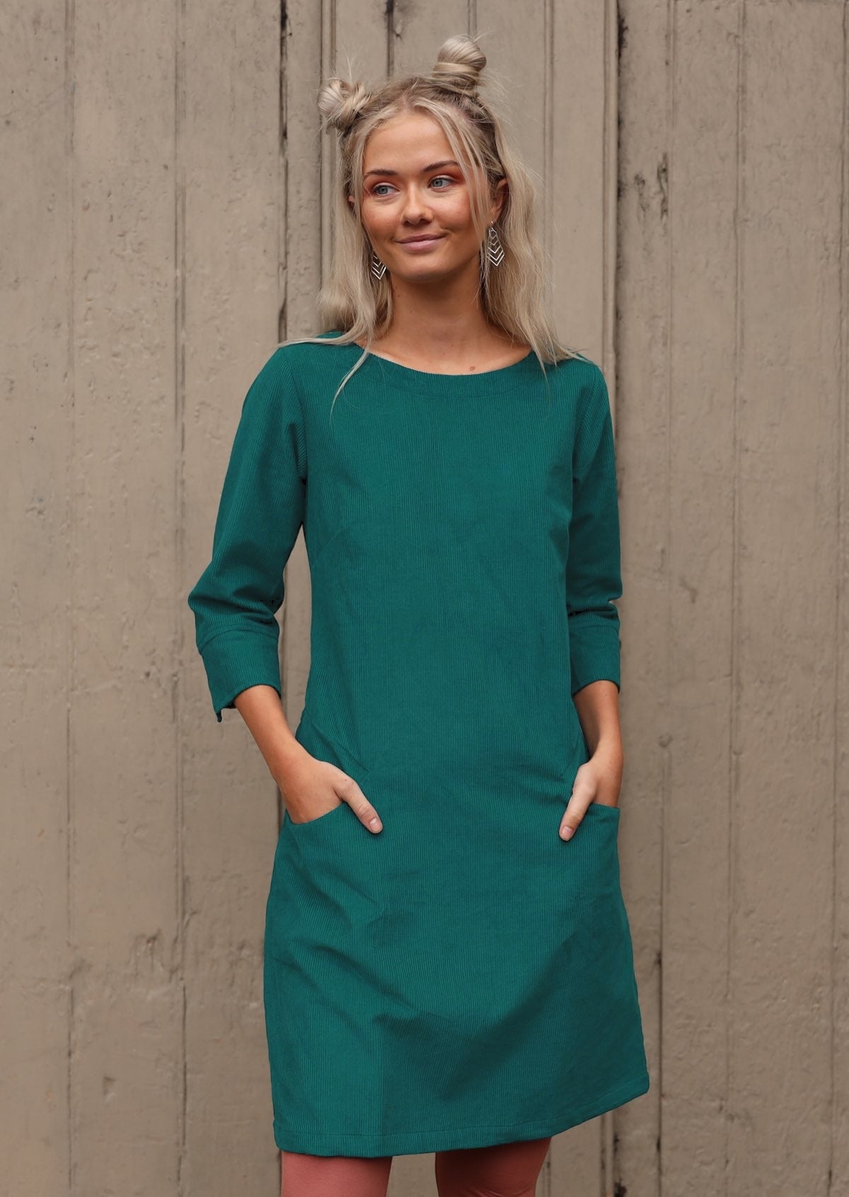 Woman with blonde hair wearing teal corduroy dress over pink leggings hands in pockets in front of old door