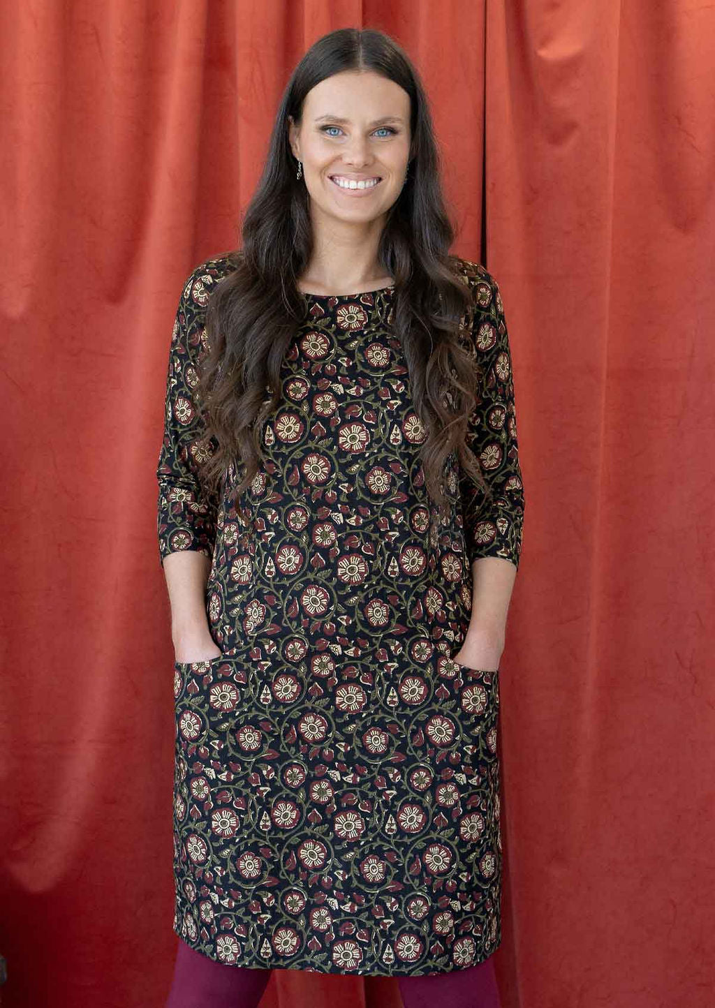 Model standing with hands in pockets wearing 100% cotton dress in cream and maroon florals on black base.