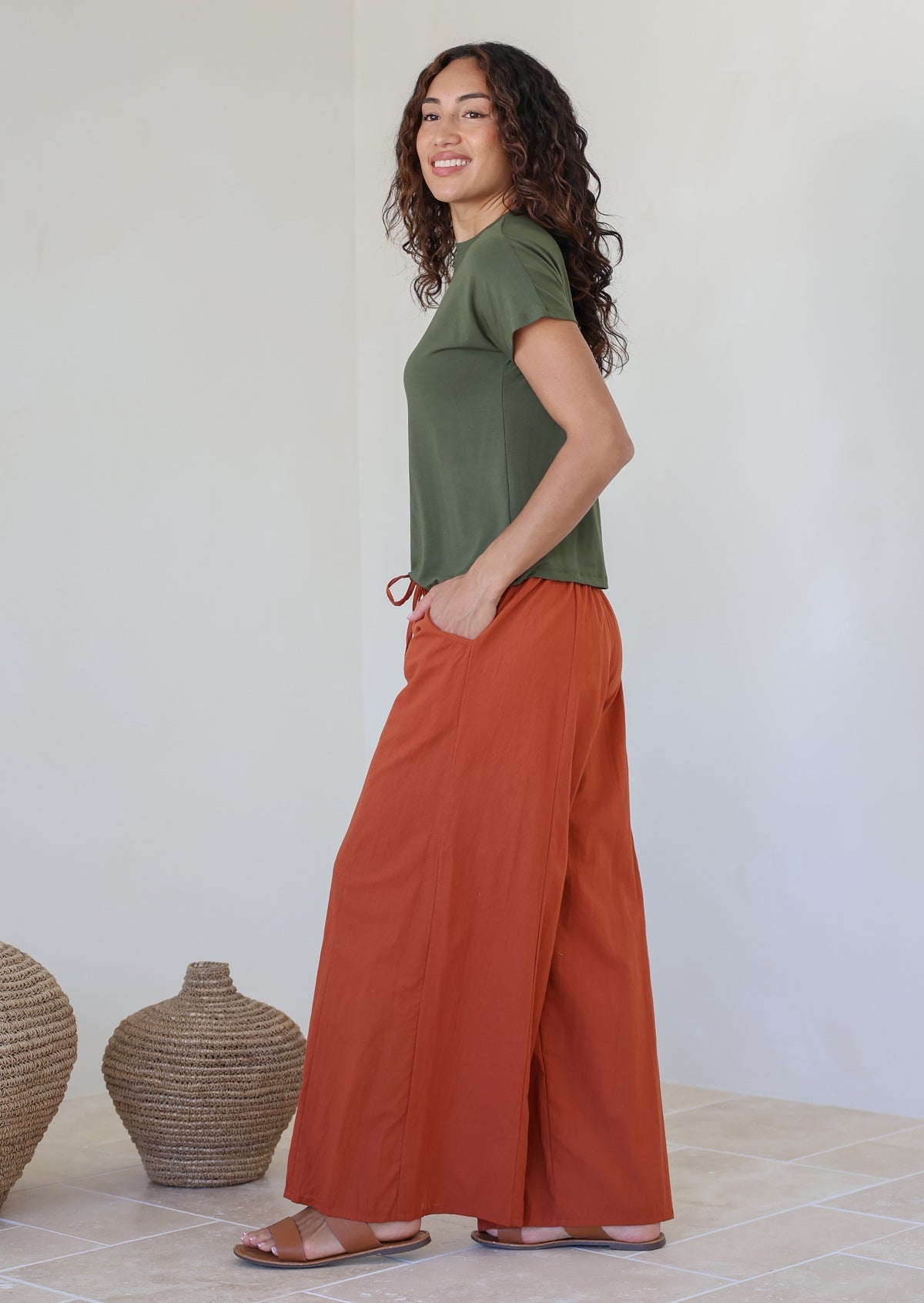 Woman posing sideways wears a green basic top and rust colored wide-leg pants standing against a white wall with hands in pockets