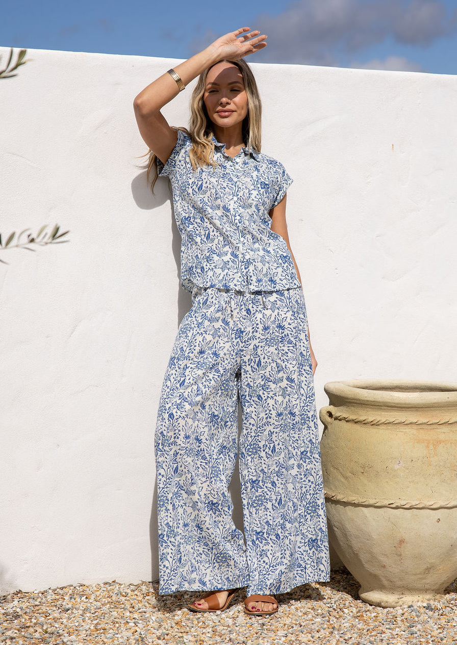 Woman in a white based blue floral cotton pant against a white wall.