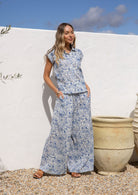 Woman in a white based blue floral cotton pant wide legged against a white wall.