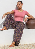 Model sitting in a relaxed pose wearing 100% cotton wide leg pants with pink, cream and light brown florals on dark purple base.