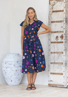 Woman wearing a midi length cotton day dress with a buttoned bodice, cap sleeves, collar and waist tie.