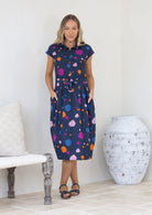 Woman wearing a navy based cotton day dress with colourful polka dots in a minimal indoor setting, with hands in pockets