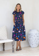 Woman wearing a blue based colourful spotty cotton dress with buttoned bodice and collar, with her hands in pockets