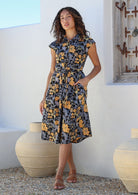 Woman wearing a midi length retro floral cotton dress with buttoned bodice, standing against a white wall and decorative vases
