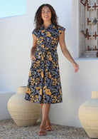 Model stands with one hand in pocket wearing a midi length floral printed shirt style cotton dress with cap sleeves, buttoned bodice and a removable waist tie