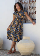 Woman leans on a wall wearing a shirt style cotton midi length dress with buttoned bodice, pockets, collar and cap sleeves