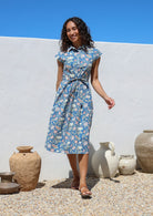 Woman in a blue floral shirt style cotton dress with buttoned bodice standing outdoors with a white wall and decorative pots in the background
