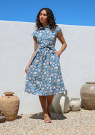 Model standing with one hand in pocket wears 100% cotton shirt style midi length dress with cap sleeves, fabric waist tie and small gathers at the waist