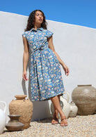Woman leans on a wall wearing a dusty blue floral printed midi length cotton dress with collar, cap sleeves, buttoned bodice and pockets