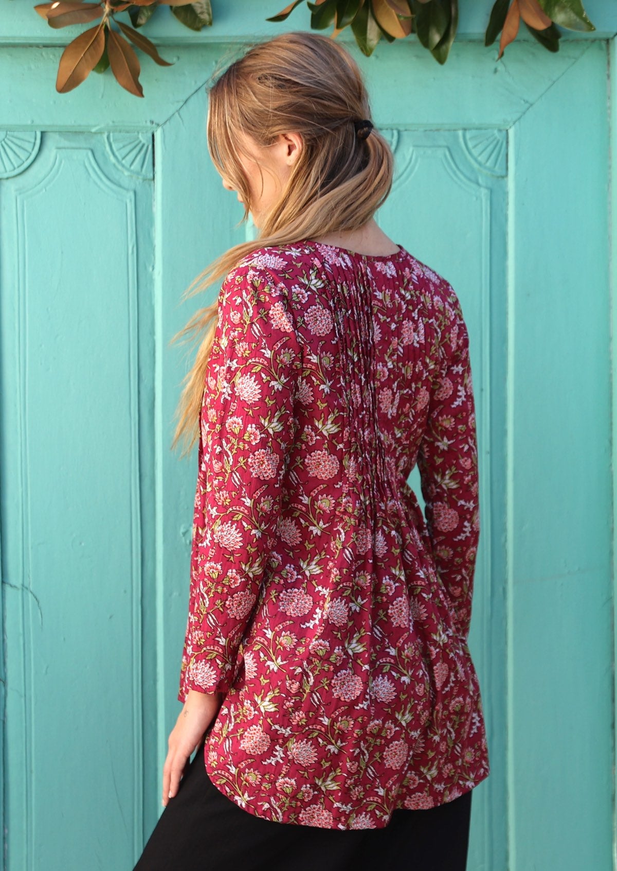 Woman shows back of cotton long sleeve top with small pin tucks across centre back that lead into flared base that covers the derrière