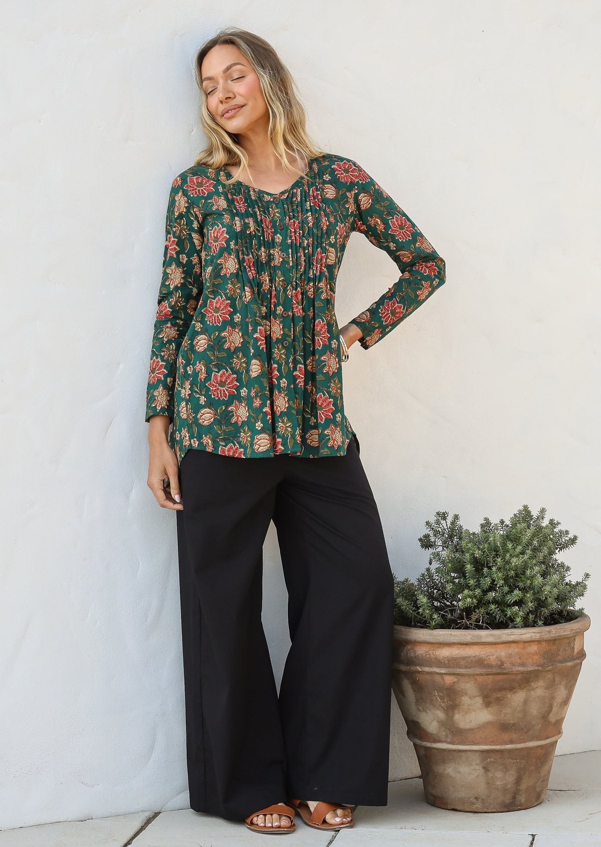Woman wearing a cotton floral long-sleeve button through shirt and black pants standing next to a potted plant.