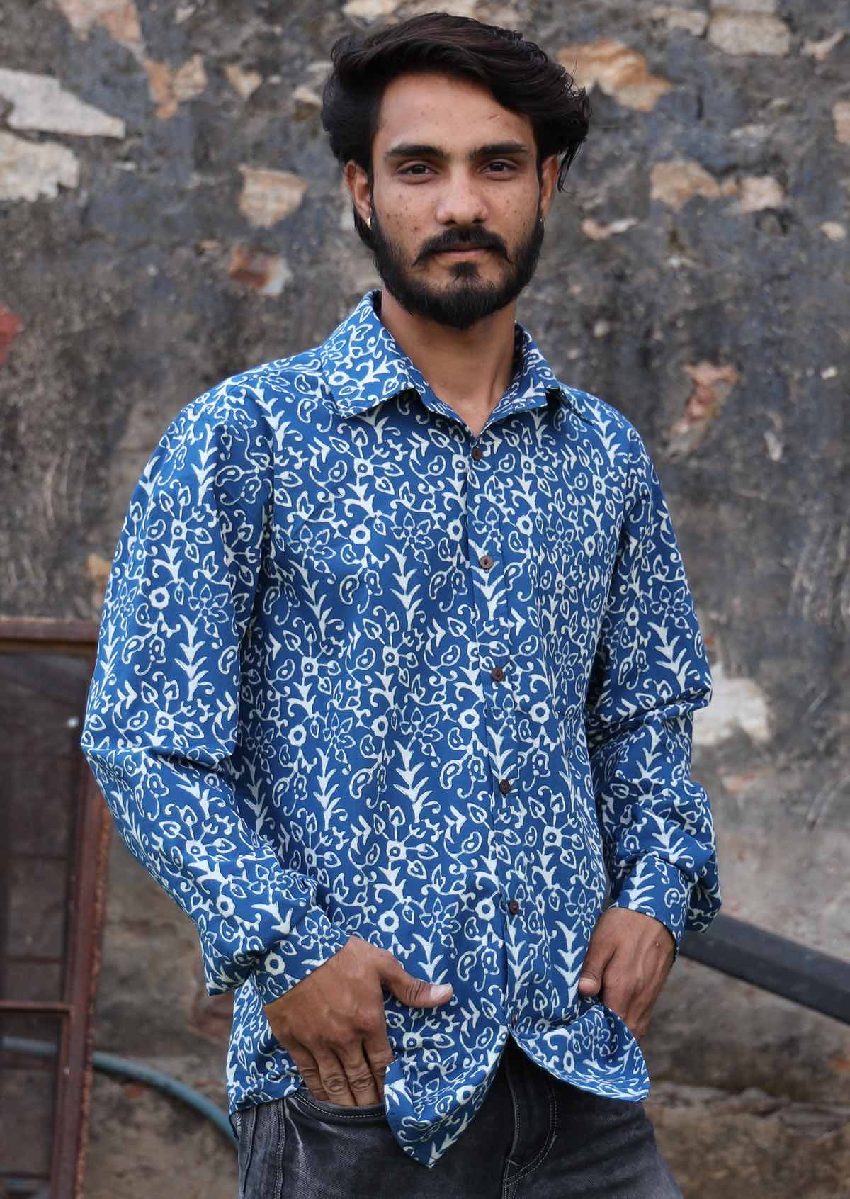 Man wears long sleeve collared shirt in blue based white floral print, with hands in pockets of pants
