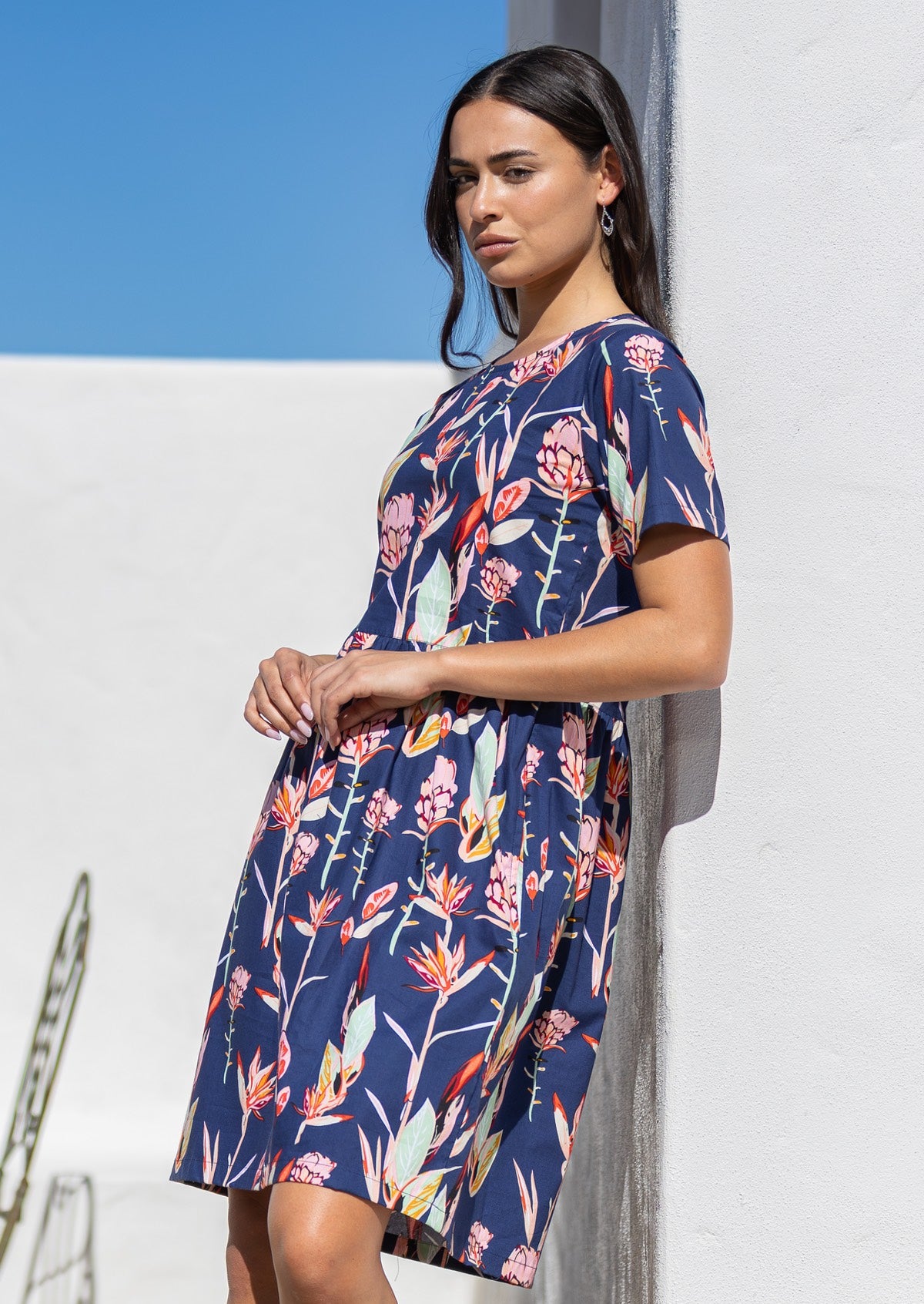 Woman leaning against the wall in dark navy blue based floral print dress with small pin tucks at the waist.