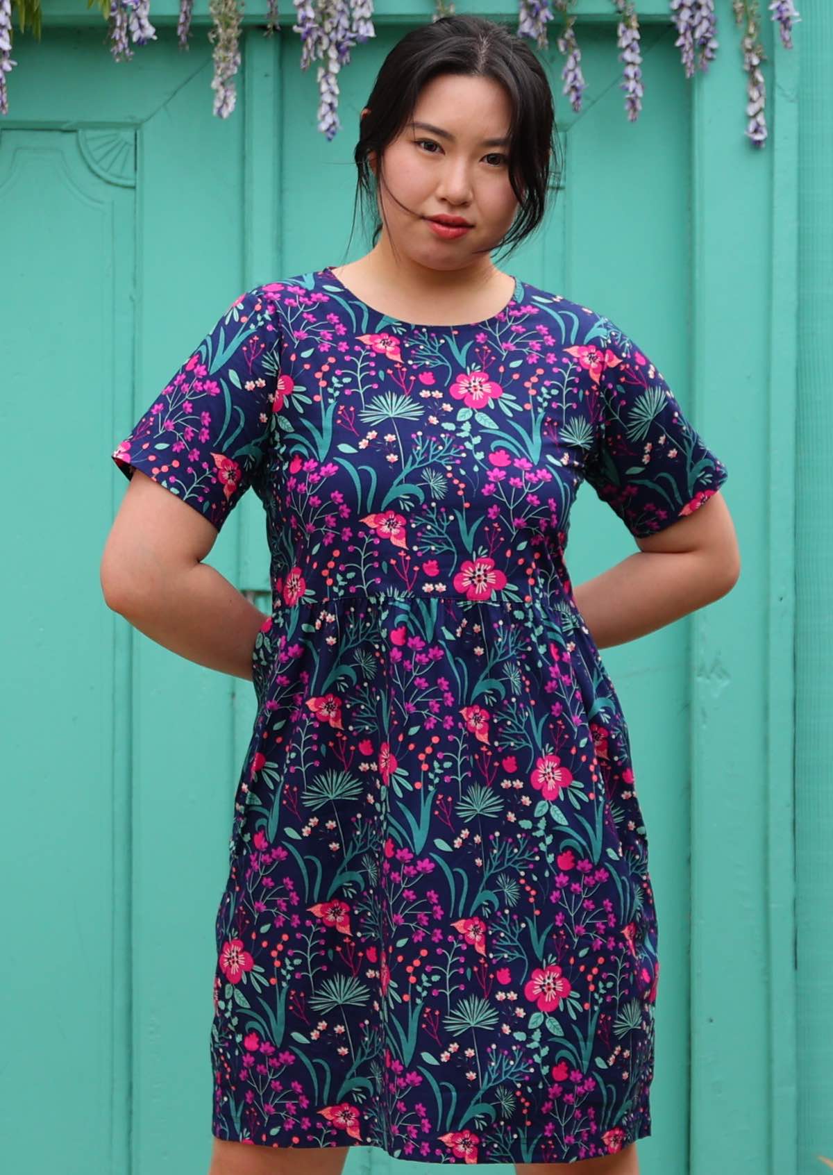 woman in loose fit purple, navy and pink floral print 100% cotton dress in front of aqua door hands behind back
