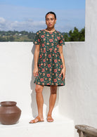 Woman wearing relaxed fit, above the knee length cotton dress with short sleeves and bright floral print on deep green base.