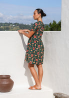 Model standing sideways wearing a loose fit, above the knee length cotton dress with pinkish florals on deep green base.