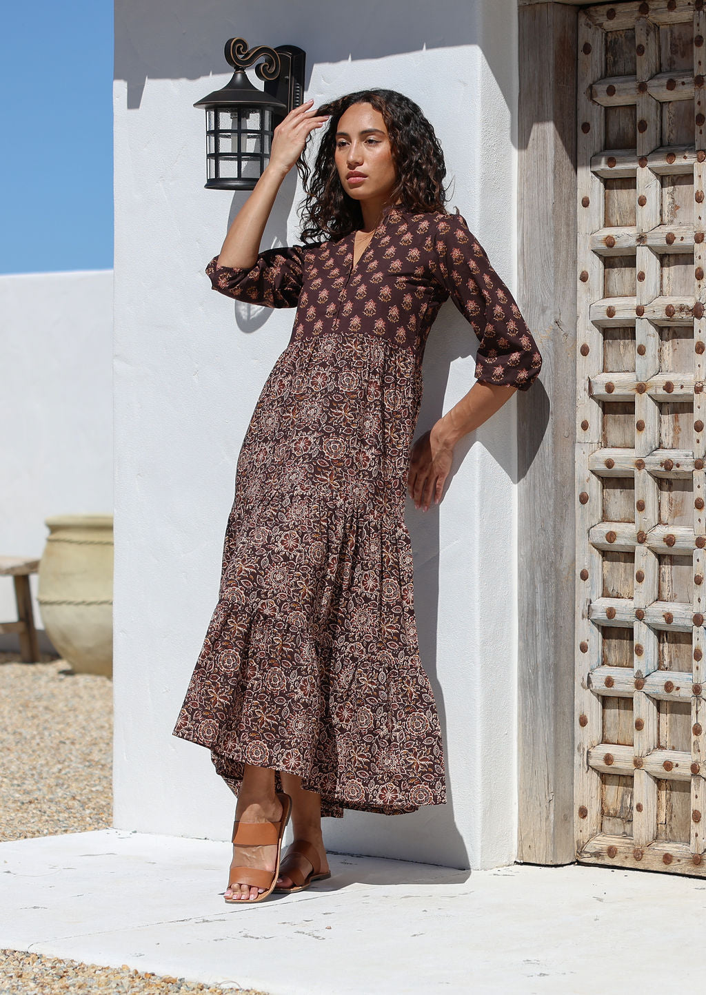 Woman in a floral cotton dress standing against a white wall with a wooden door.