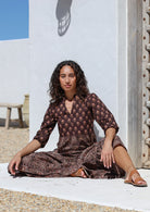 Woman in a Indian cotton maxi dress sitting on the ground with a white wall and blue sky in the background.