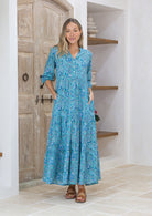 Woman wearing a blue floral maxi dress with 3/4 sleeves, buttoned bodice and hands in pocket.