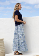 Woman wearing a white based blue floral cotton skirt standing against a white wall.