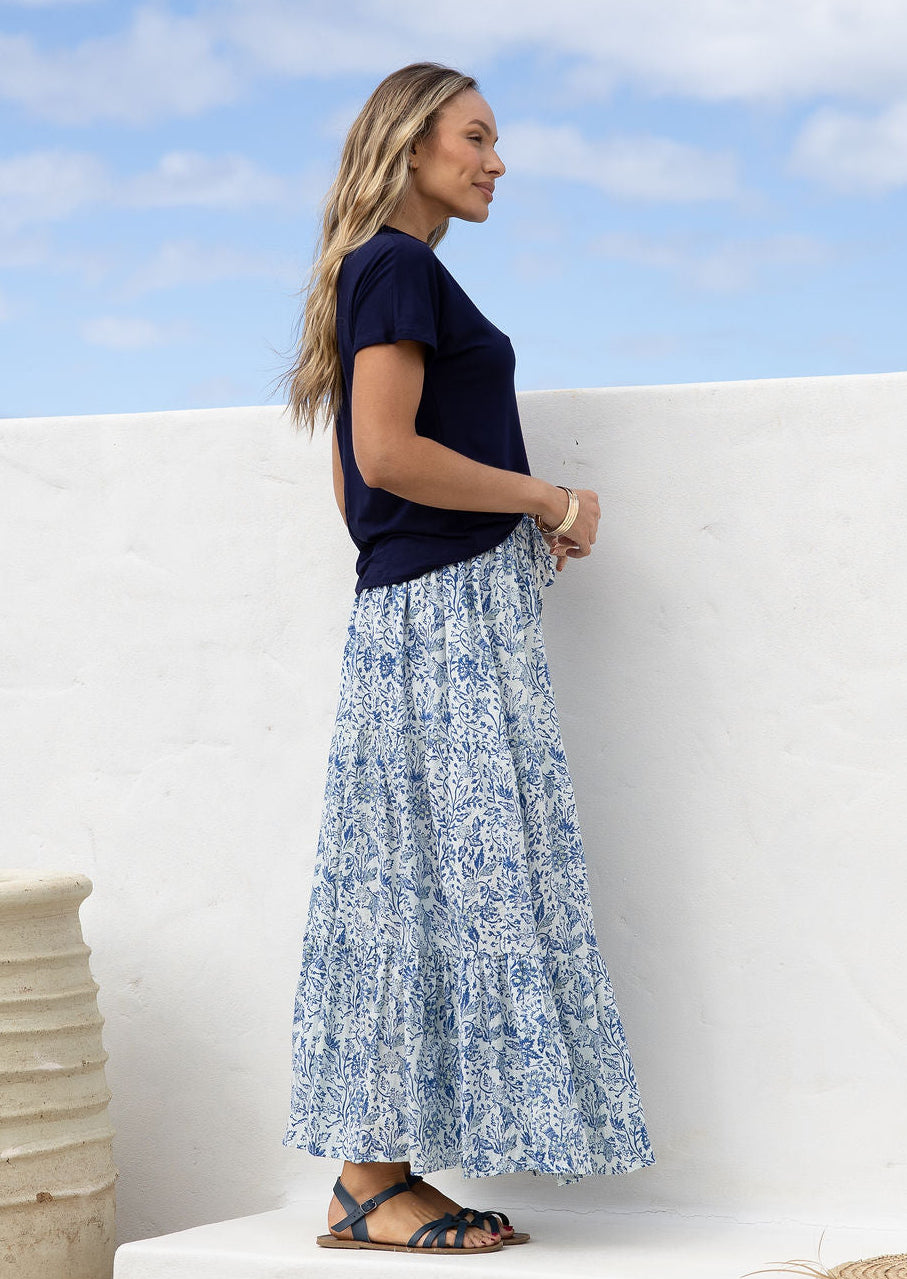 Woman wearing a white based blue floral cotton skirt standing against a white wall.