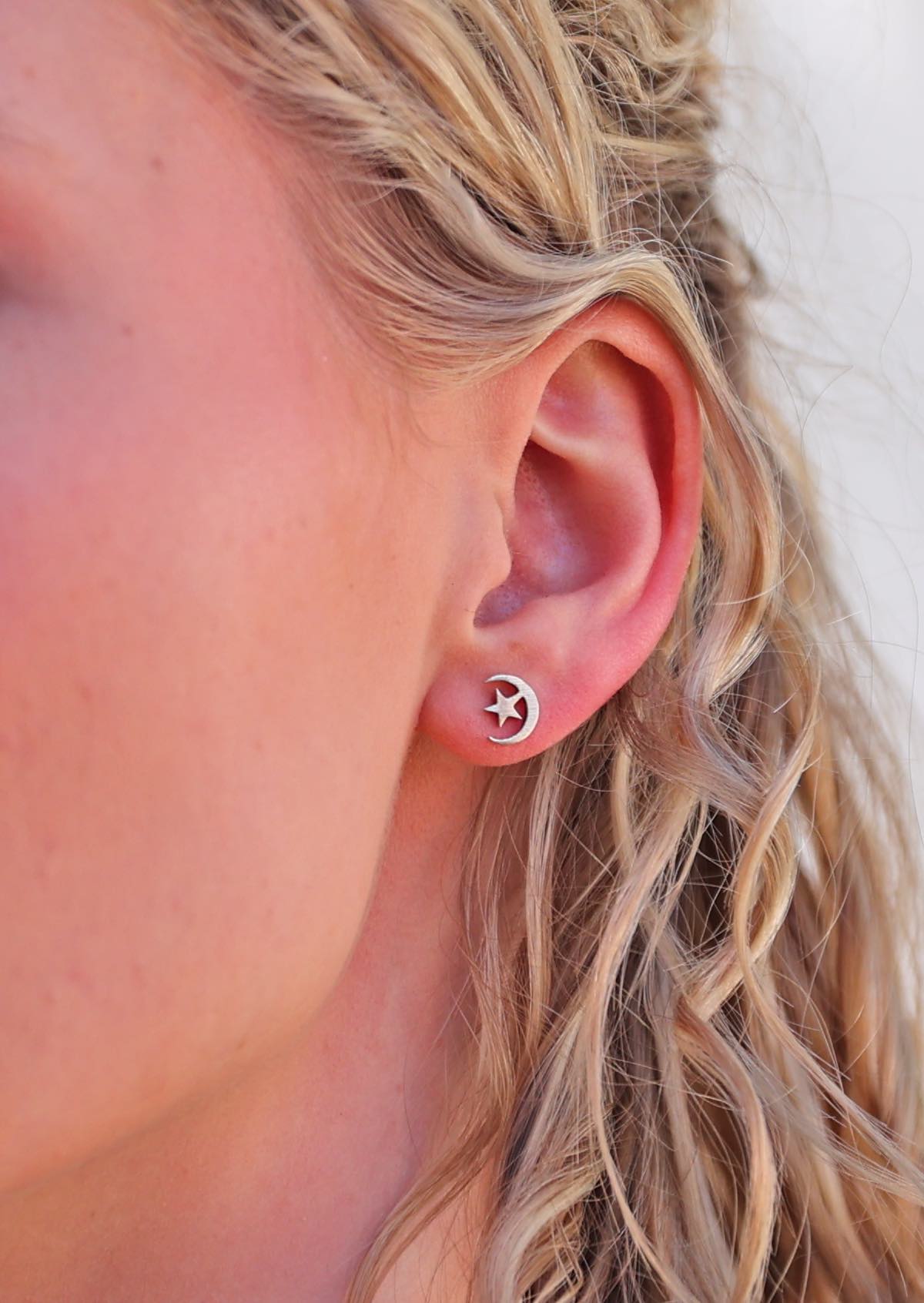 A close-up image of a woman wearing Moon and Stars silver stud earrings shaped like a crescent moon with a star inside it.