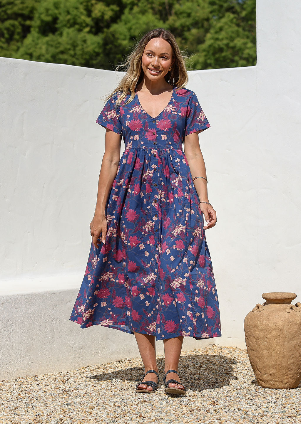 Woman standing in a 100% cotton floral print dress in blue with a V-neckline, short sleeves, pockets, and buttons through to the knee.