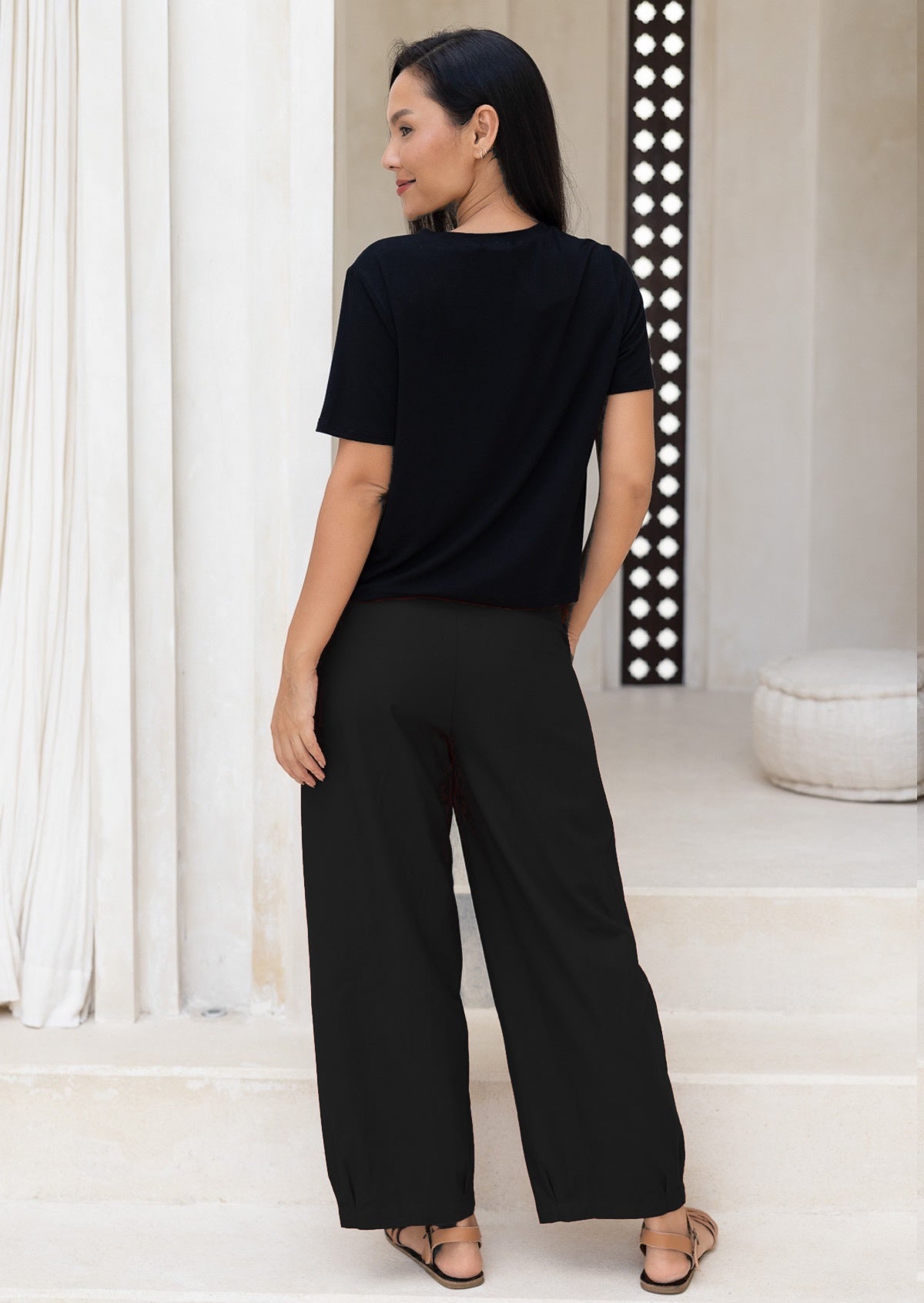 Woman wearing a black cotton pants and t-shirt standing in a room with white walls and a decorative wall.