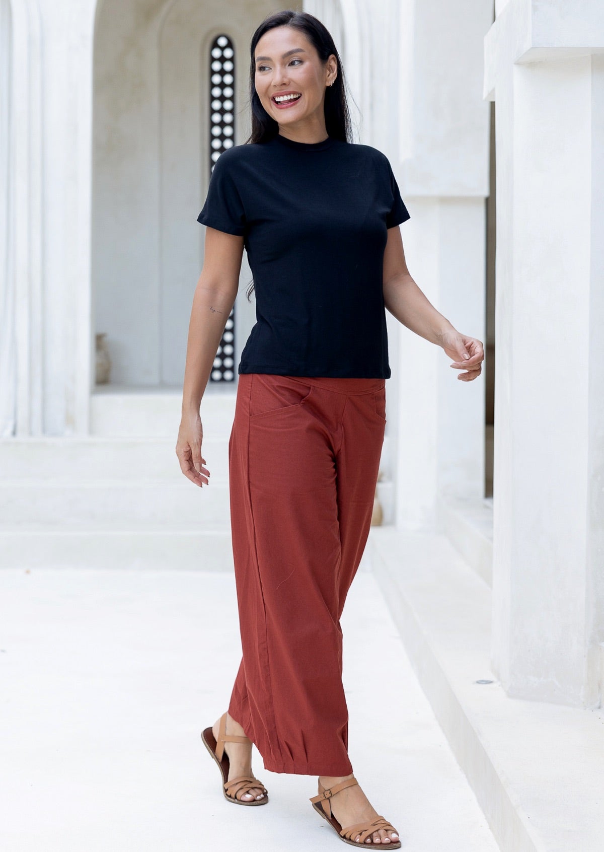 Woman wearing rust-colored cotton pants with pockets and pleated ankles, standing in a bright open area.