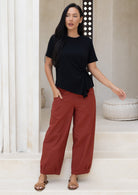 Model wears rust colour wide leg cotton pants with hand in pocket paired with basic black rayon top