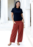 Woman with hands in pocket wears 100% cotton wide leg pants in rust with tapered ankles, side zipper and pleated ankles