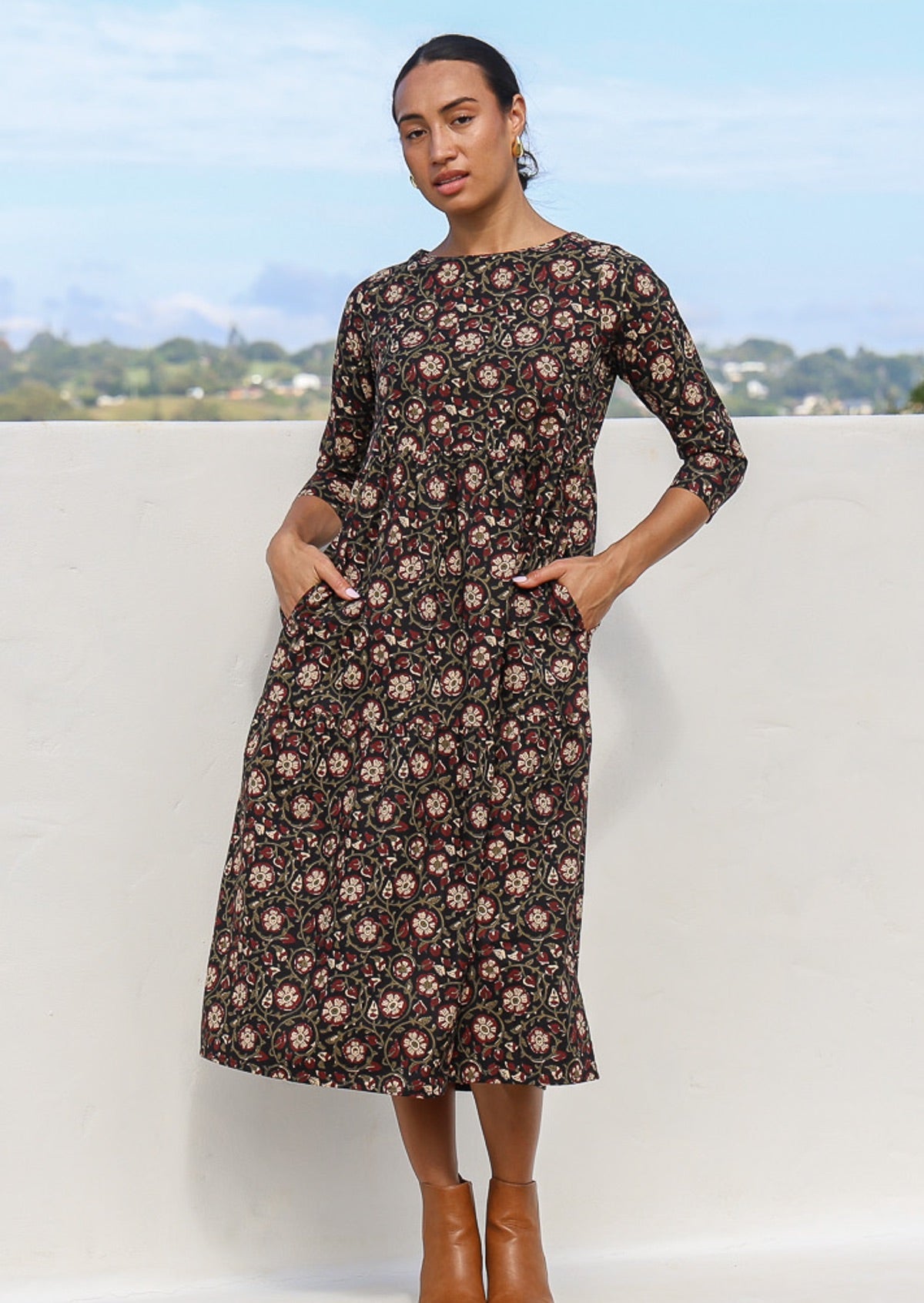 Model with hands in pocket wearing a relaxed fit black cotton dress with maroon, cream and olive florals.