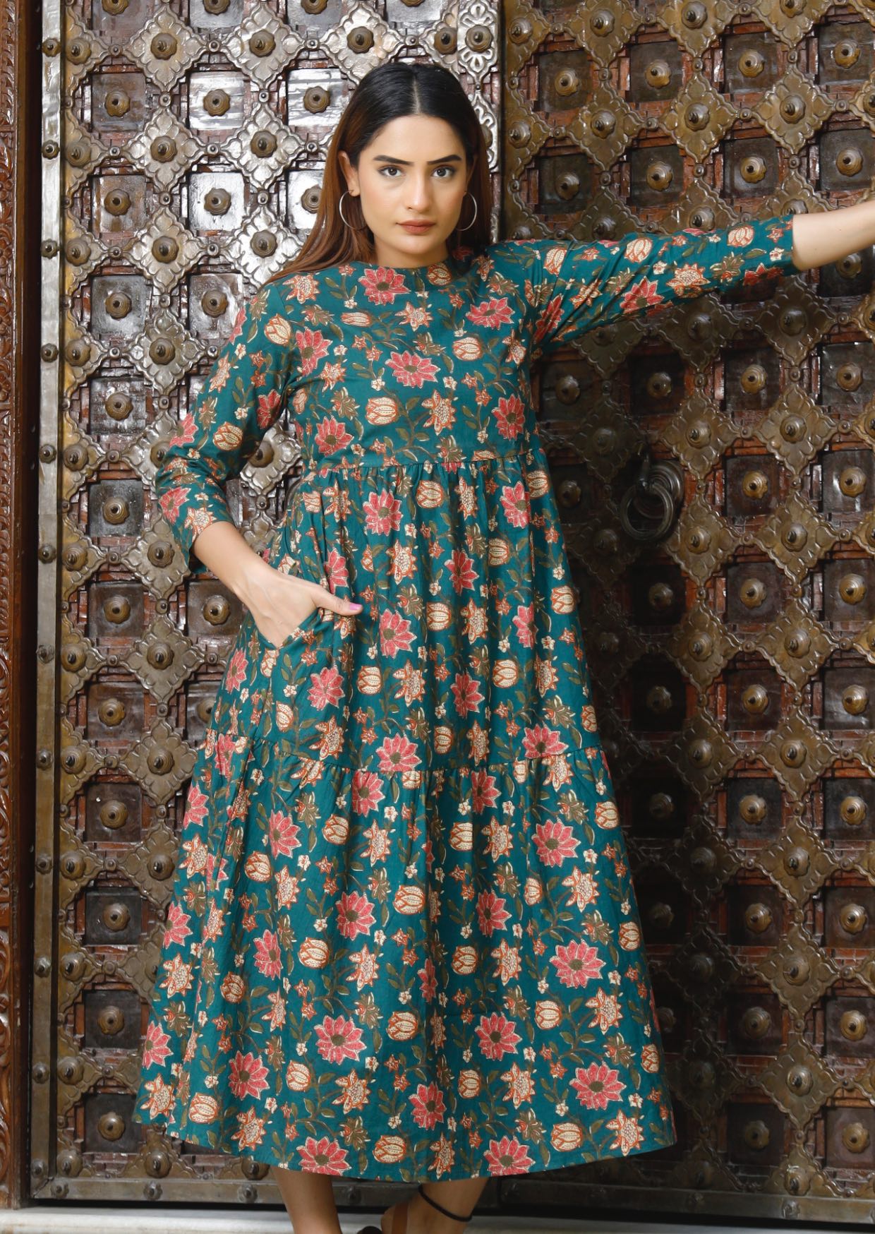 Model with dark hairs wears 100% cotton, 3 tiered midi dress in green based floral print with hand in pocket.