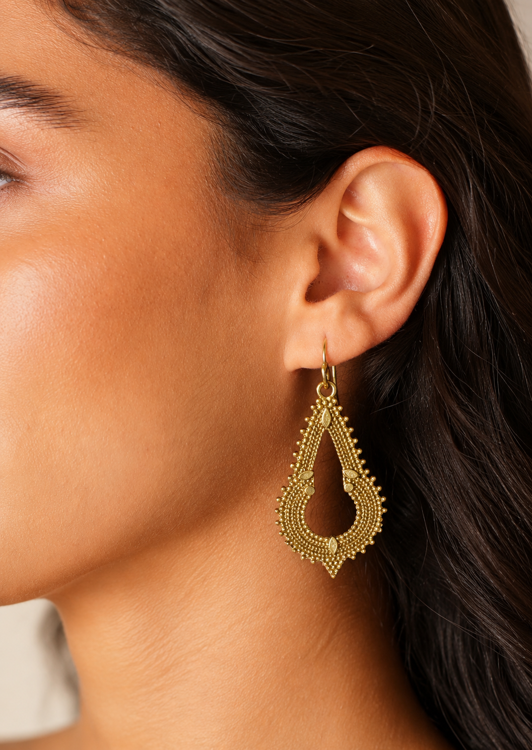 Close up of a woman wearing an elegant gold coloured teardrop shaped earring that has layered rope like detailing, tiny raised bead accents, and small leaf motifs inspired by traditional Indian jewellery
