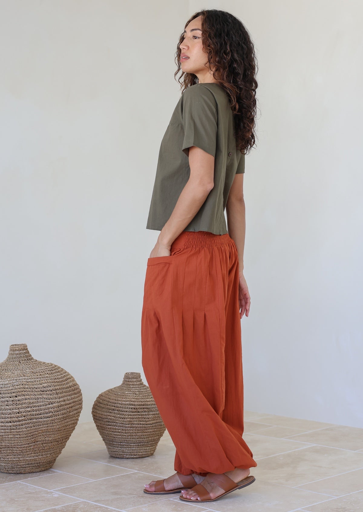 Woman wearing a rust-colored pants with hand in pockets standing against a white background