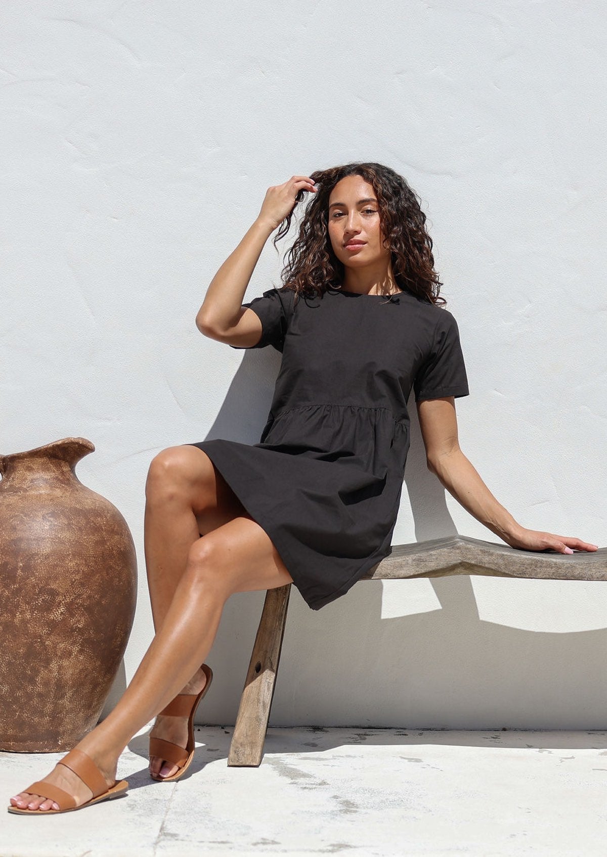 Woman in a above knee length black cotton dress sitting on a wooden bench against a white wall