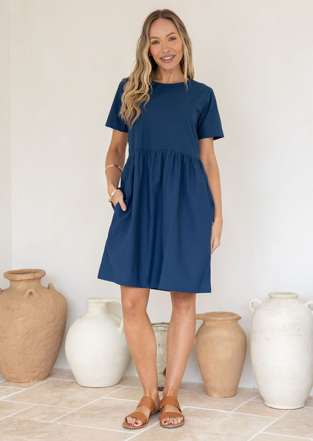 Model wears sea blue cotton dress with length above the knee, pockets and small gathers at the loose waist