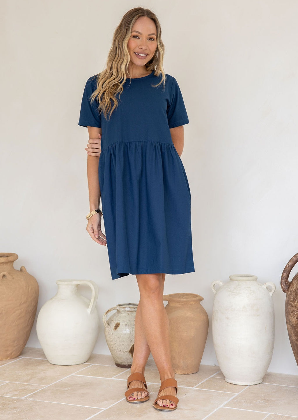 Woman standing in a blue cotton dress with short sleeves, relaxed fit and pockets, standing in front of decorative vases
