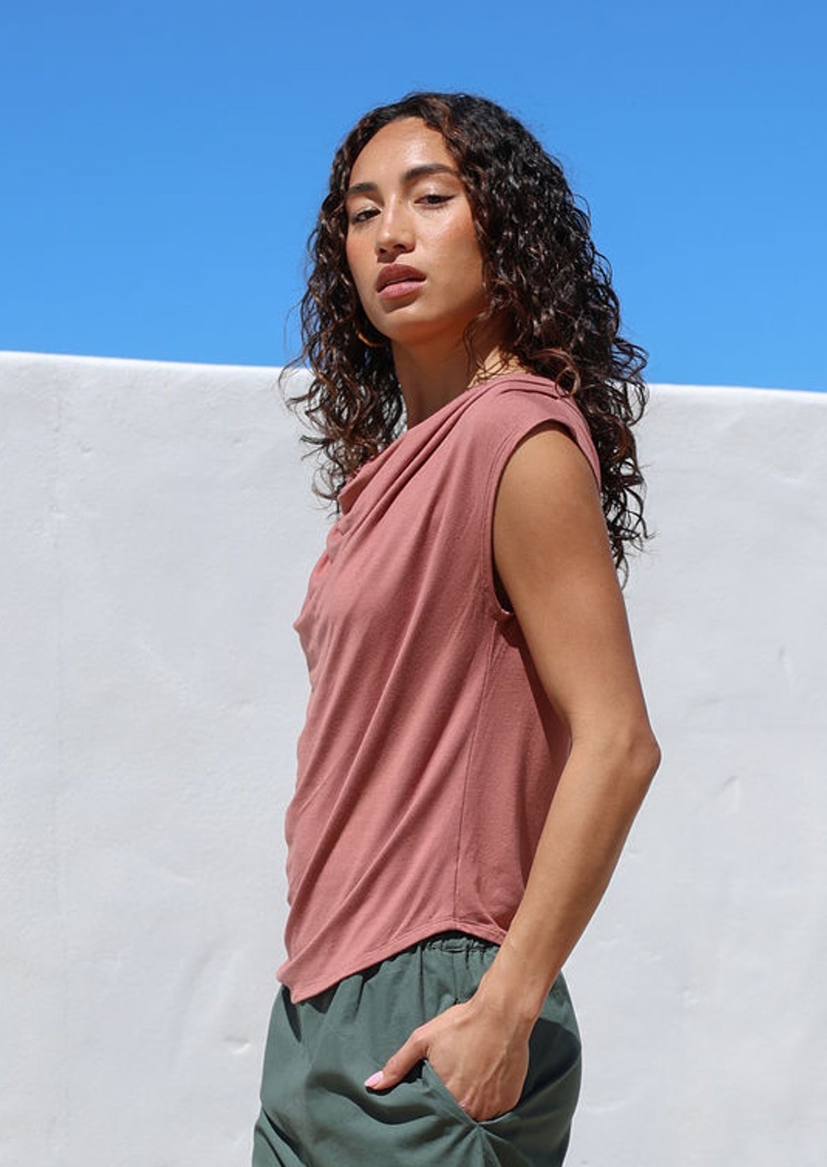 Woman wearing a pink sleeveless top with draped neckline against a white wall with blue sky.